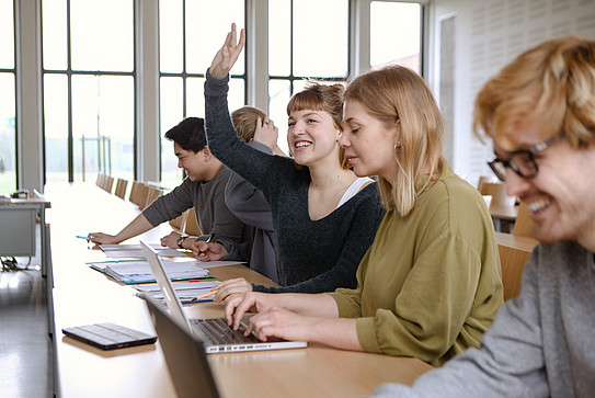Five students sit in the seminar room with their laptops, one of them give a hand signal.