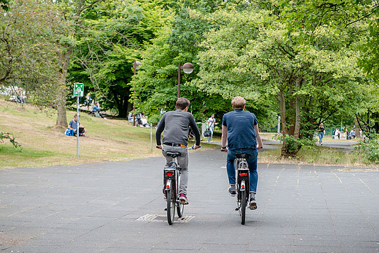Foto: Zwei Männer fahren auf dem grünen Campus der RUB Fahrrad.