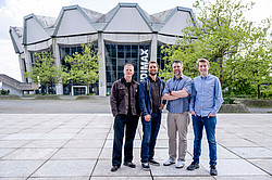 Photo: Four men smile at the camera. In the background: the main lecture hall at Ruhr University Bochum.