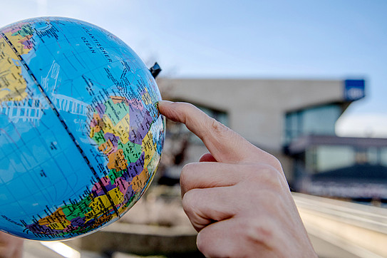 Finger points to a spot on a globe, with the campus of Ruhr University Bochum in the background