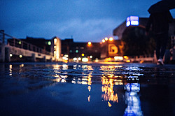 Foto: Eine Wasserpfütze bei Dunkelheit. In ihrer Reflektion spiegeln sich die Beleuchtung und Gebäude auf dem Campus der Ruhr Universität Bochum.
