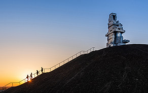 Side view of a slag heap, groups of people climbing up or down stairs, on the slag heap there is a sculpture made of stones