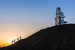 Side view of a slag heap, groups of people climbing up or down stairs, on the slag heap there is a sculpture made of stones