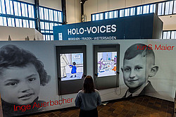 Photo: A person looks at interactive screens with illustrations in an exhibition featuring the sign “HOLO-VOICES.”
