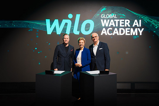 Auf dem Foto sieht man Oliver Hermes, Ina Brandes und Rektor Prof. Manfred Bayer vor einer digitalen Leinwand stehen. Sie stehen nebeneinander und lächeln für das Foto in die Kamera.