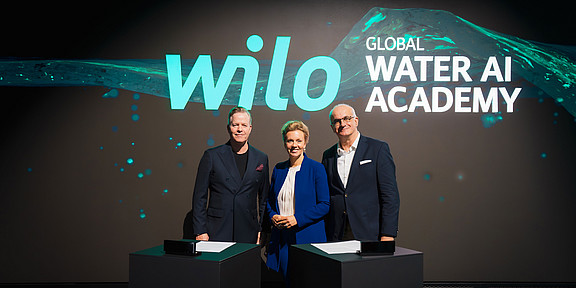 In the photo, Oliver Hermes, Ina Brandes, and President Prof. Manfred Bayer are standing in front of a digital screen. They are standing side by side and smiling at the camera for the photo.