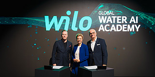 In the photo, Oliver Hermes, Ina Brandes, and President Prof. Manfred Bayer are standing in front of a digital screen. They are standing side by side and smiling at the camera for the photo.